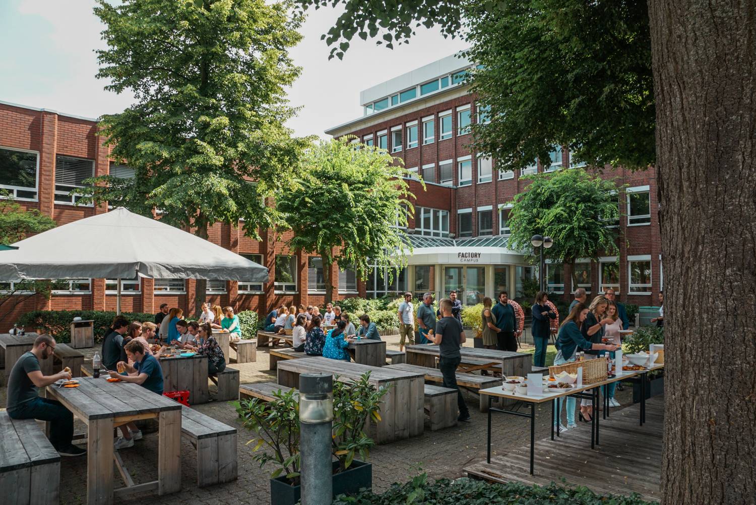 Der Außenbereich des Factory Campus mit Menschen, die an Holztischen sitzen und Essen verspeisen.